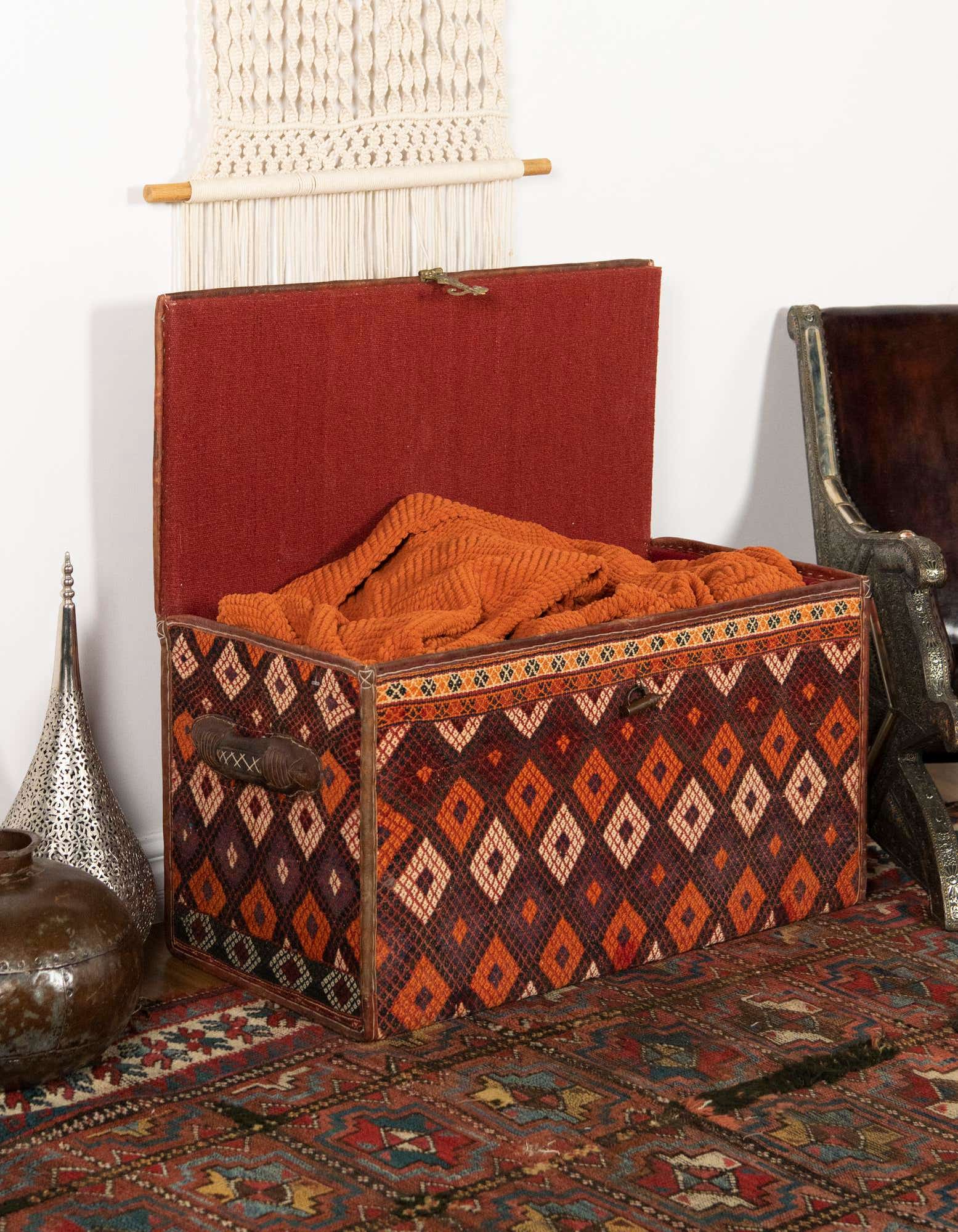 A 2x3 rectangular traditional rug with a rich blend of orange, red, and blue geometric patterns, grounding a decorative storage trunk.