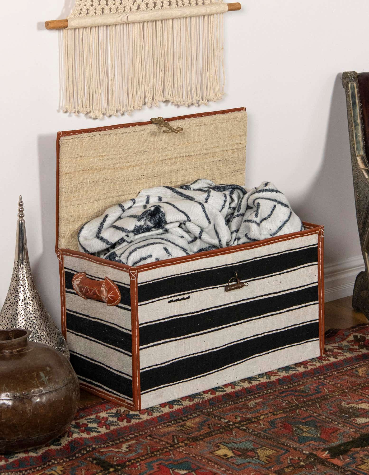 A 1x2 rectangular black and ivory striped storage trunk with a partially open lid, sitting on a textured, handmade traditional oriental rug in a living room.