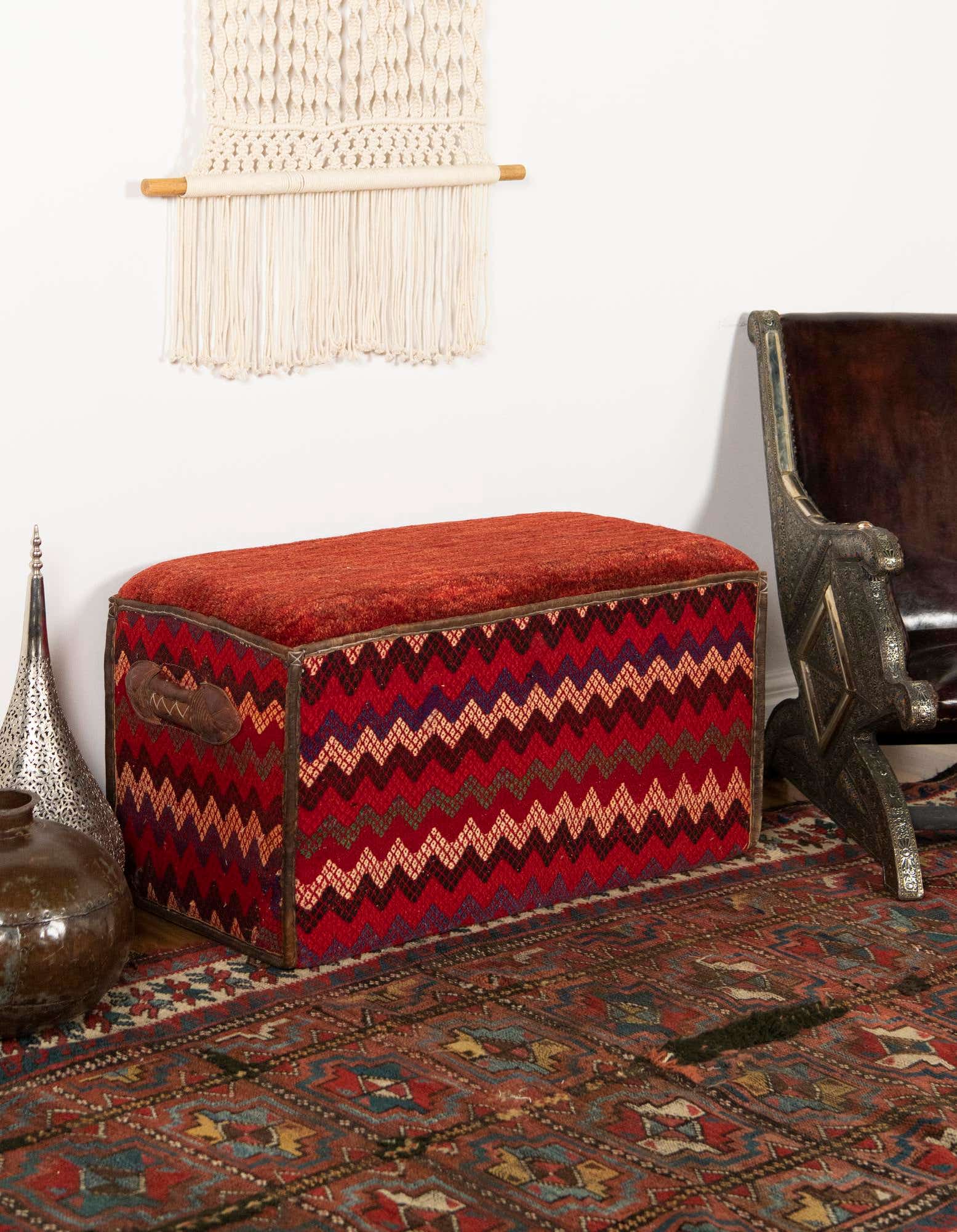 A 2x3 rectangular orange and red traditional rug with intricate patterns on a wooden floor, next to a patterned storage ottoman.