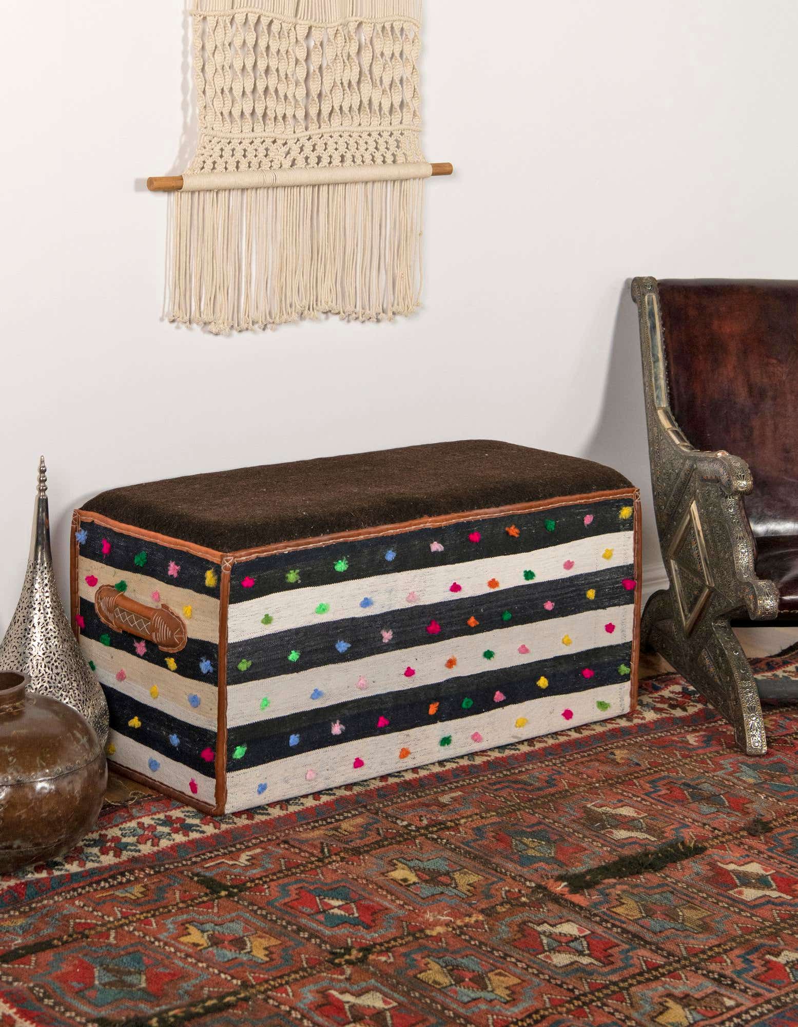 A rectangular brown and tan rug with intricate traditional patterns in red, blue, and gold, placed under a striped storage ottoman.