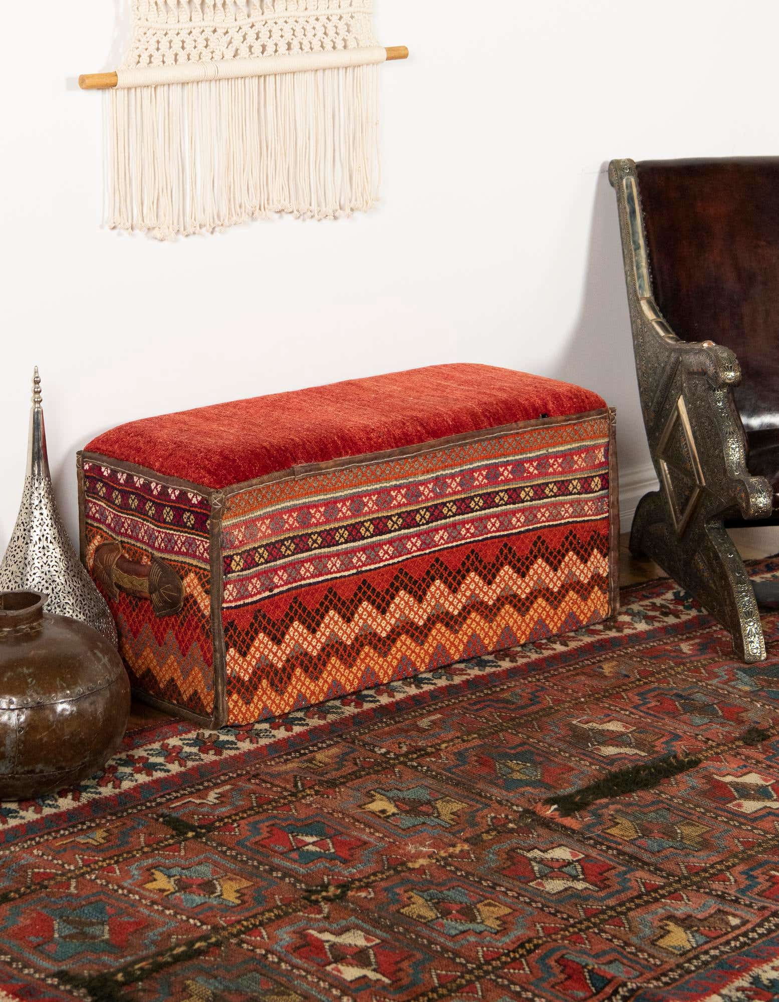 A 2x3 rectangular orange and red traditional rug with intricate patterns, placed under a colorful storage bench in an indoor setting.