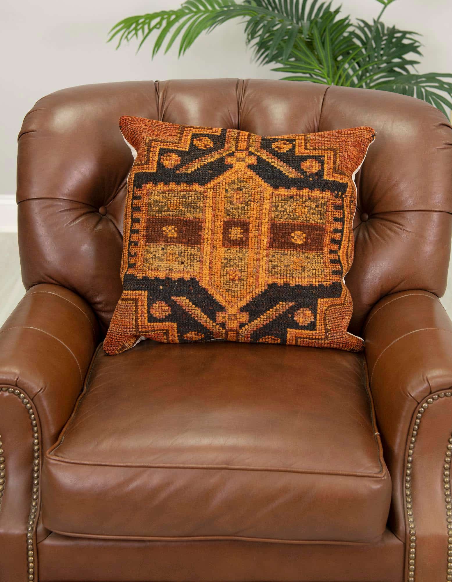 A rectangular 25 Euro pillow with a vibrant orange and red Persian pattern, made of 100% wool, resting on a brown leather armchair.