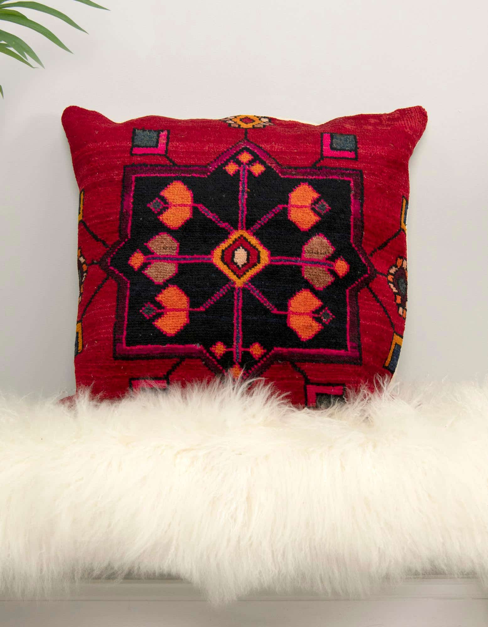 A red Persian wool pillow with a black geometric pattern and orange accents, resting on a white fluffy rug.