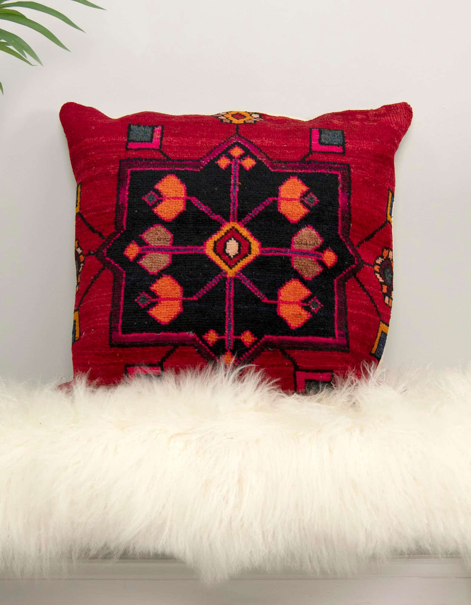 A red Persian wool pillow with a black geometric pattern and orange accents, resting on a white fluffy rug.