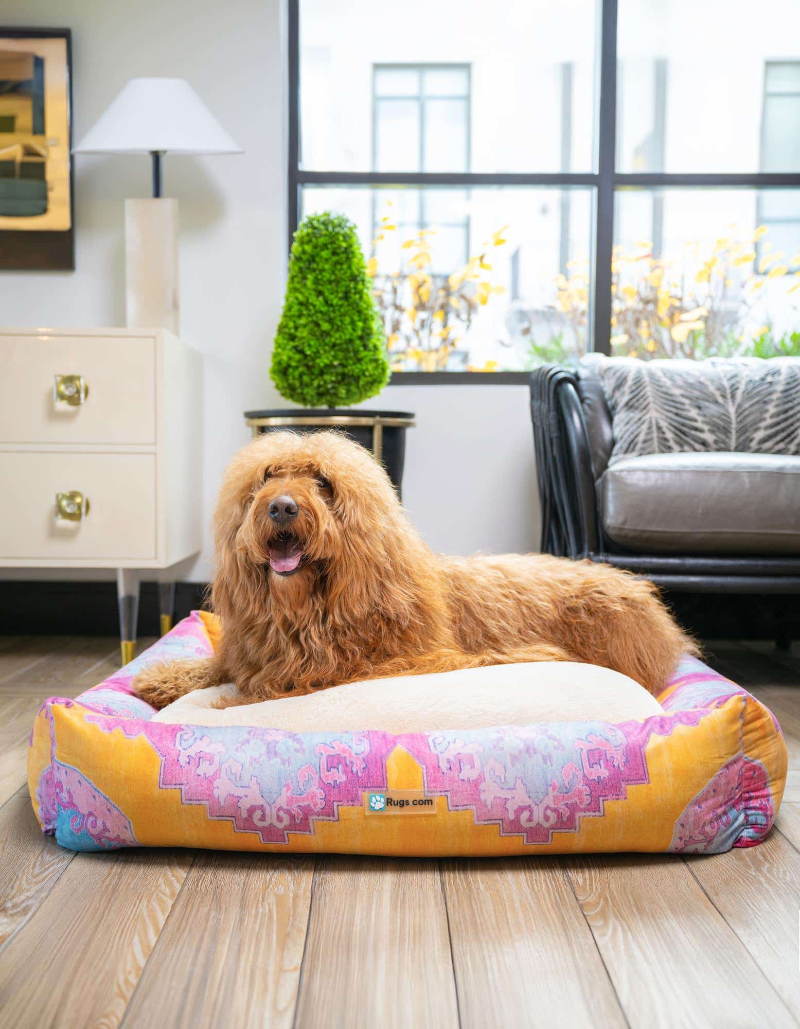 A fluffy golden dog rests comfortably on a multi-colored rectangular pet bed with a vibrant geometric pattern, set on a light wood floor indoors.