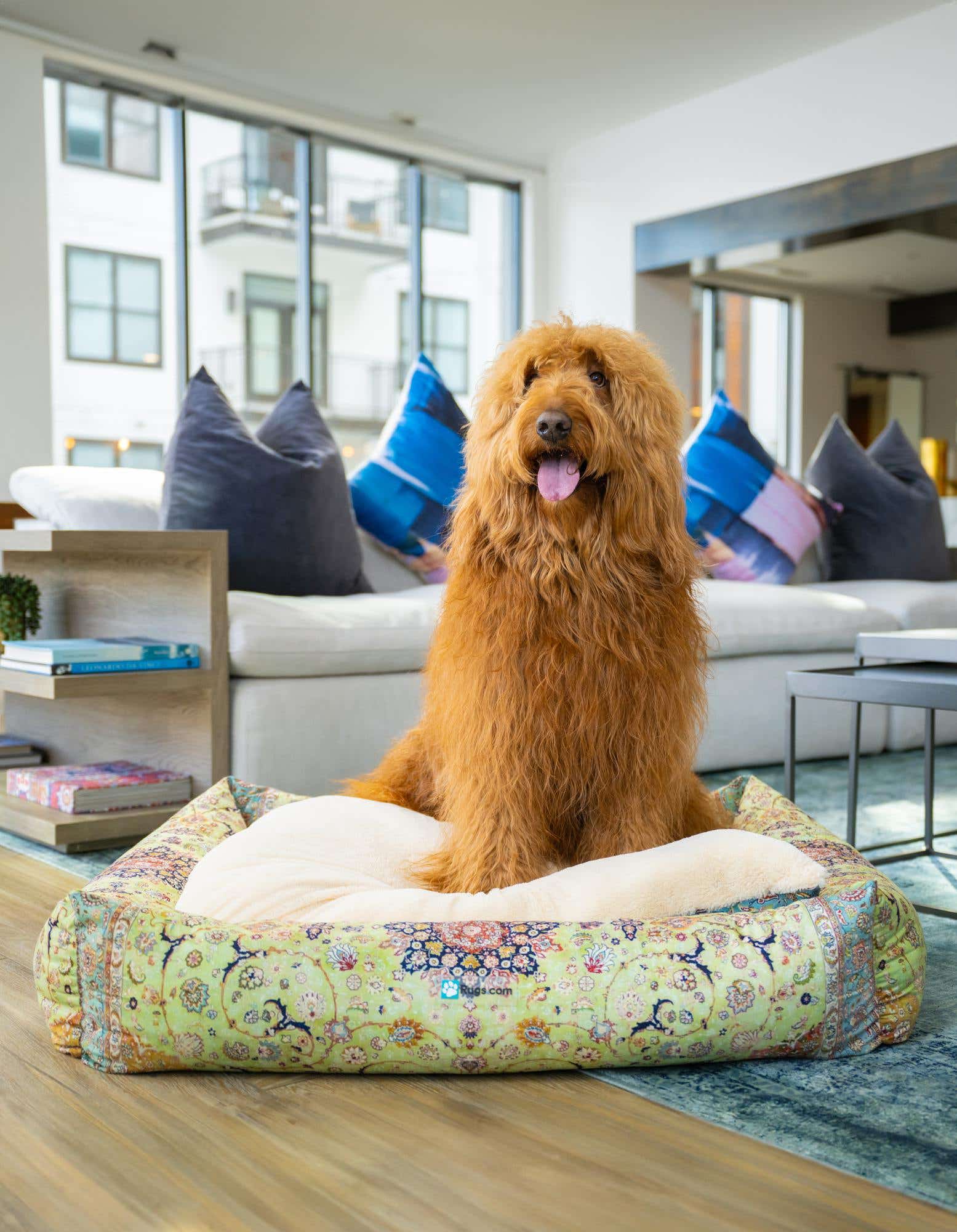 A fluffy golden doodle dog sits in a rectangular pet bed with a multi-colored traditional pattern, on a wooden floor.