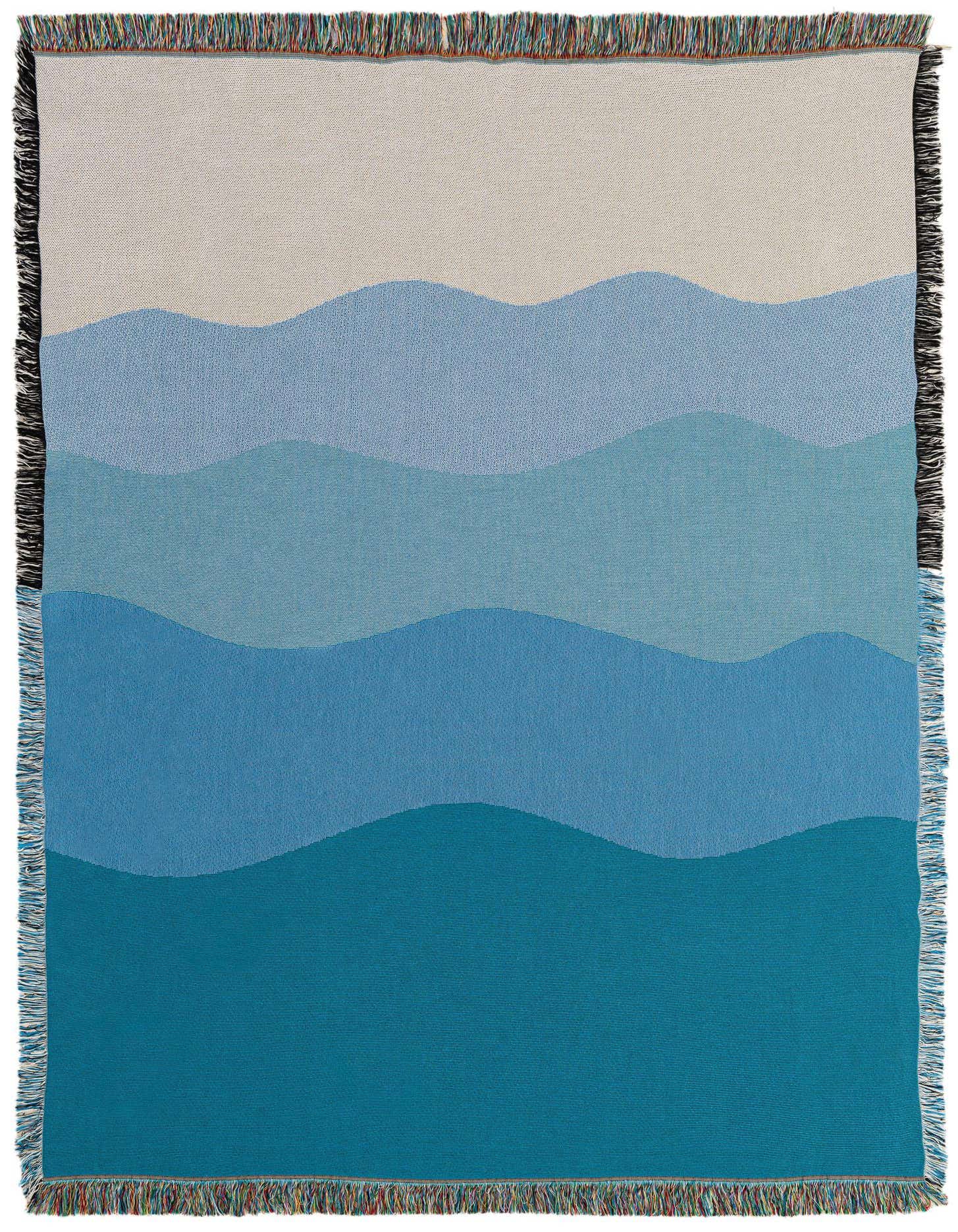 A 4x6 rectangular throw blanket with a modern abstract design featuring wavy bands of varying blue shades, from light sky blue to deep teal, with a light beige top band, and fringed edges.