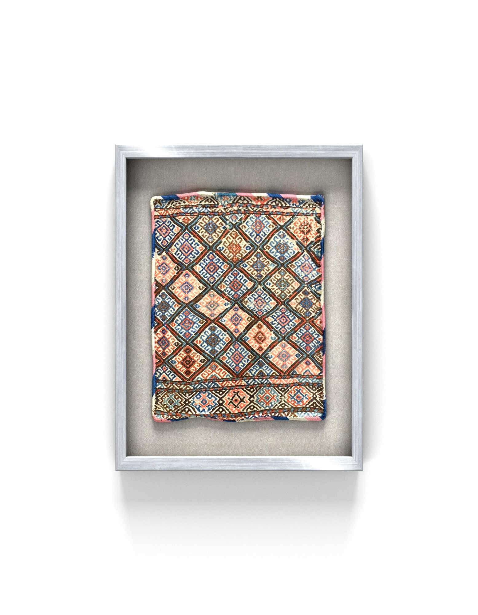 A square, multi-colored traditional textile wall art piece with intricate geometric patterns, framed in a light silver frame.